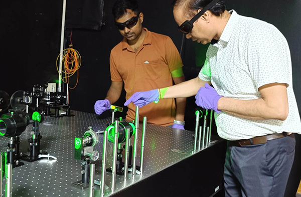 Two researchers wearing protective laser safety glasses and purple gloves align optical components emitting green light on an optics table in a laboratory.